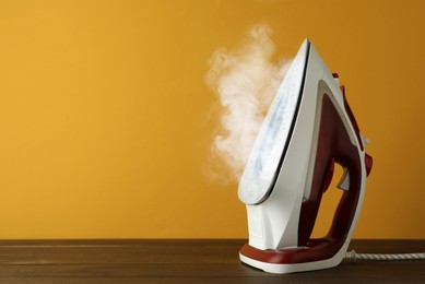 Modern iron with steam on wooden table against orange background, space for text