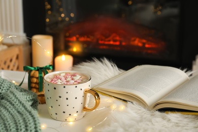 Cup of cocoa, book and cookies near fireplace indoors