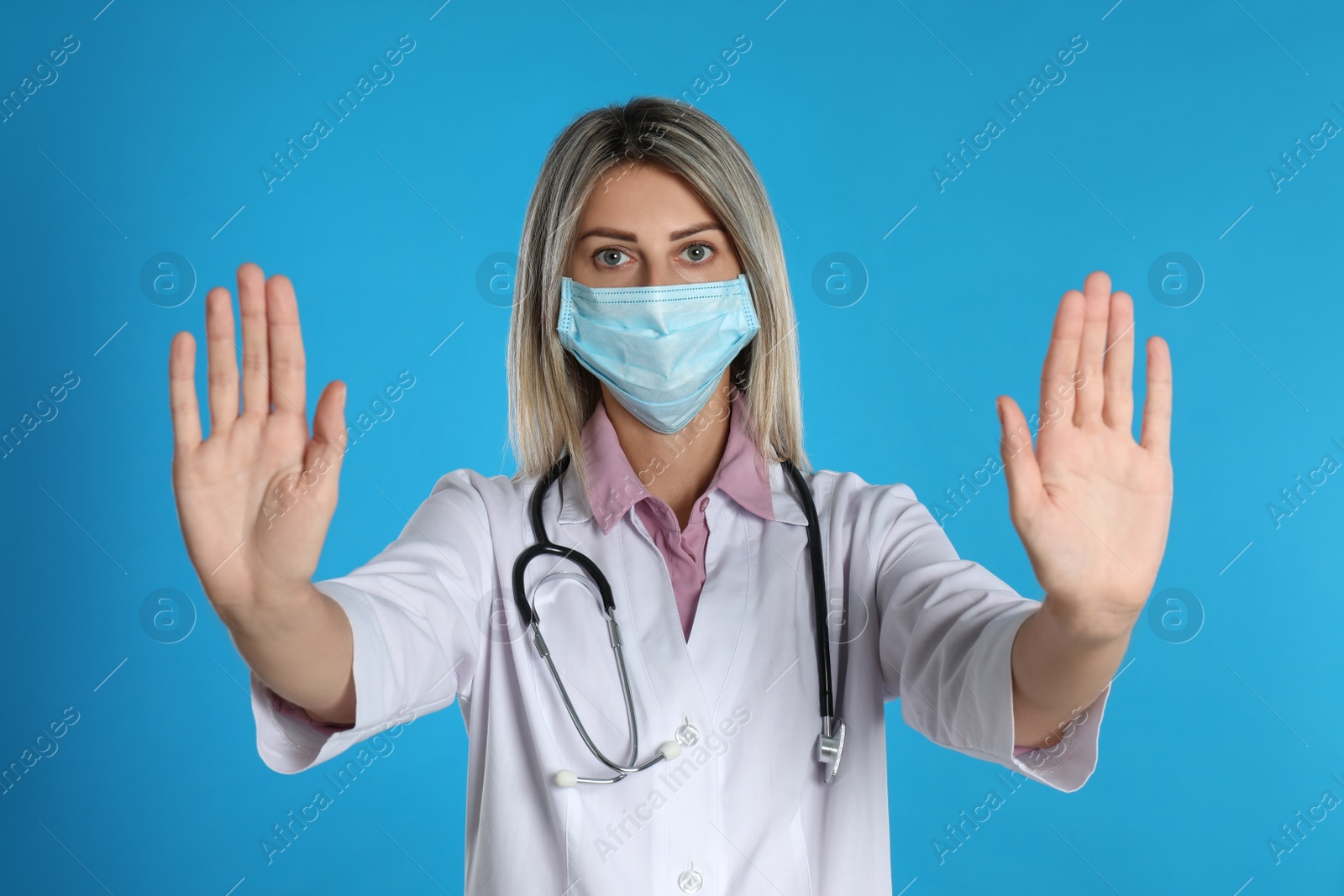 Photo of Doctor in protective mask showing stop gesture on light blue background. Prevent spreading of coronavirus