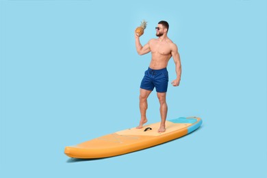 Photo of Happy man with pineapple posing on SUP board against light blue background