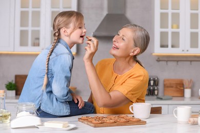 Happy grandmother feeding her granddaughter with cookie in kitchen