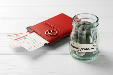 Photo of Glass jar with word Honeymoon, dollar banknotes, passports, plane tickets and golden rings on white wooden table, closeup