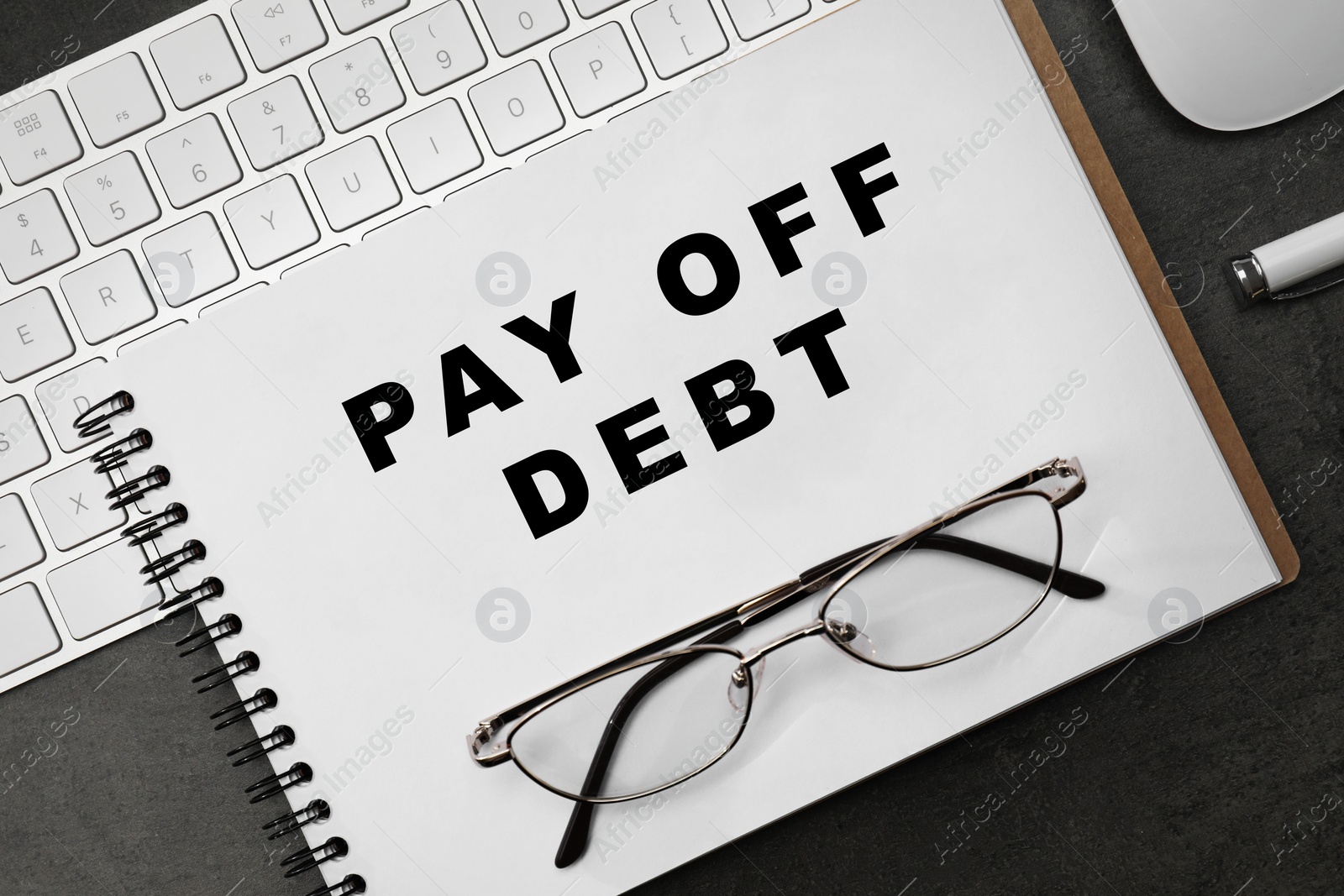 Image of Notebook with phrase Pay Off Debt, keyboard and glasses on dark table, flat lay