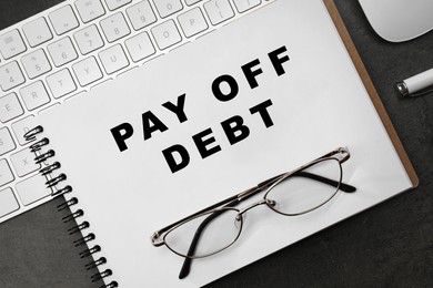 Notebook with phrase Pay Off Debt, keyboard and glasses on dark table, flat lay