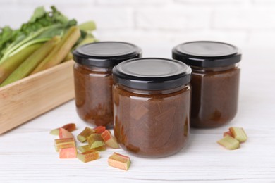 Photo of Jars of tasty rhubarb jam and cut stalks on white wooden table, closeup