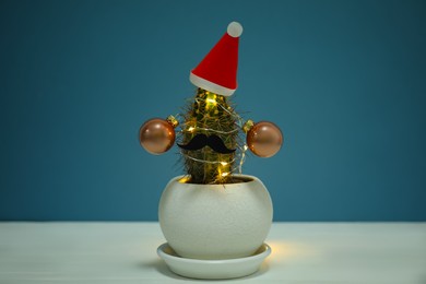 Photo of Cactus decorated with glowing fairy lights and santa hat on white table against blue background