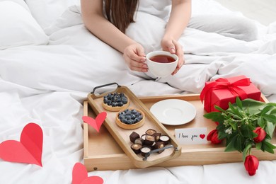 Photo of Tasty breakfast served in bed. Woman with tea, desserts, gift box, flowers and I Love You card at home, closeup