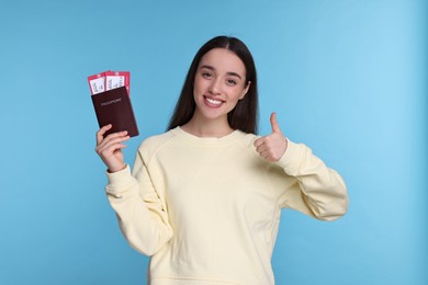 Happy woman with passport and tickets showing thumb up on light blue background