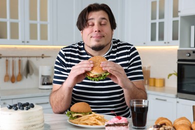 Photo of Happy overweight man with tasty burger at table in kitchen