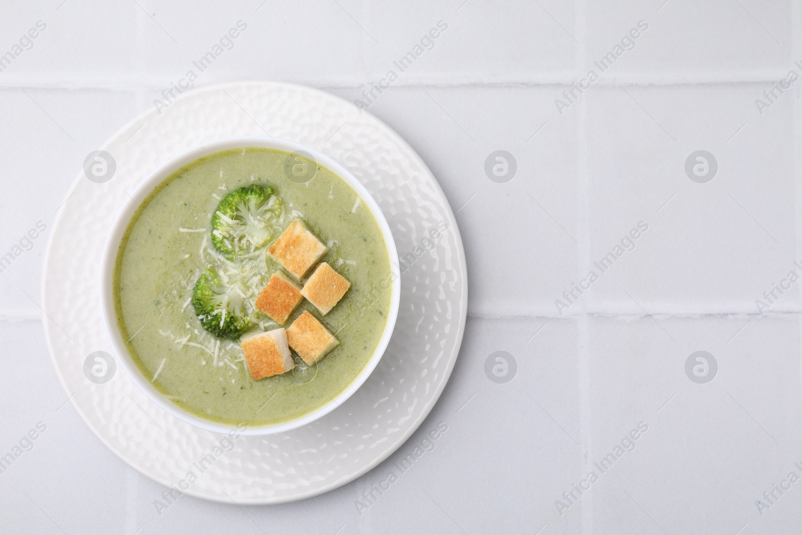 Photo of Delicious broccoli cream soup with croutons and cheese on white tiled table, top view. Space for text