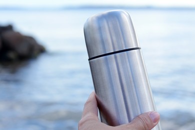 Photo of Woman holding metallic thermos with hot drink near sea, closeup. Space for text