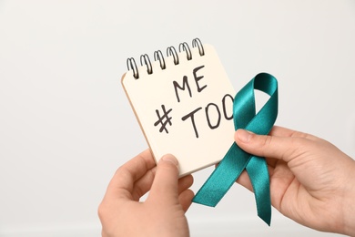 Photo of Woman holding notebook with hashtag MeToo and teal awareness ribbon against light background, closeup. Stop sexual assault