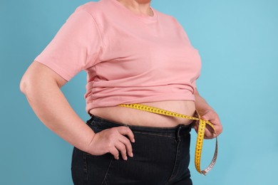 Overweight woman measuring waist with tape on light blue background, closeup