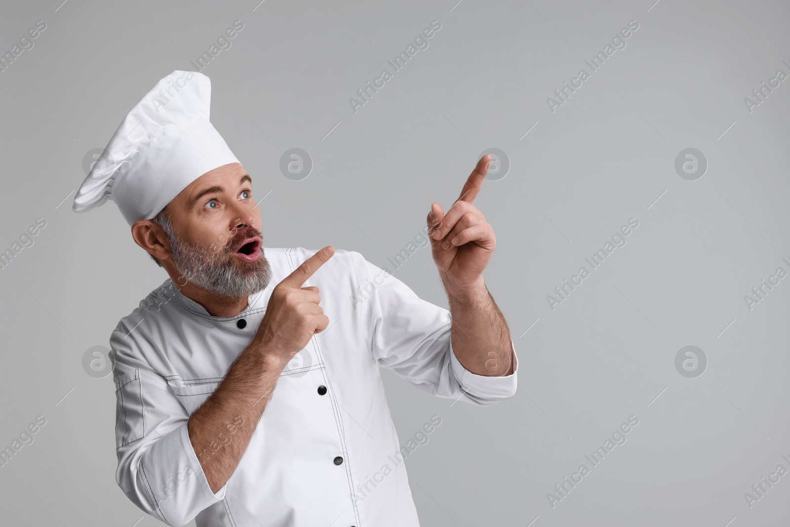 Photo of Surprised chef in uniform pointing at something on grey background, space for text