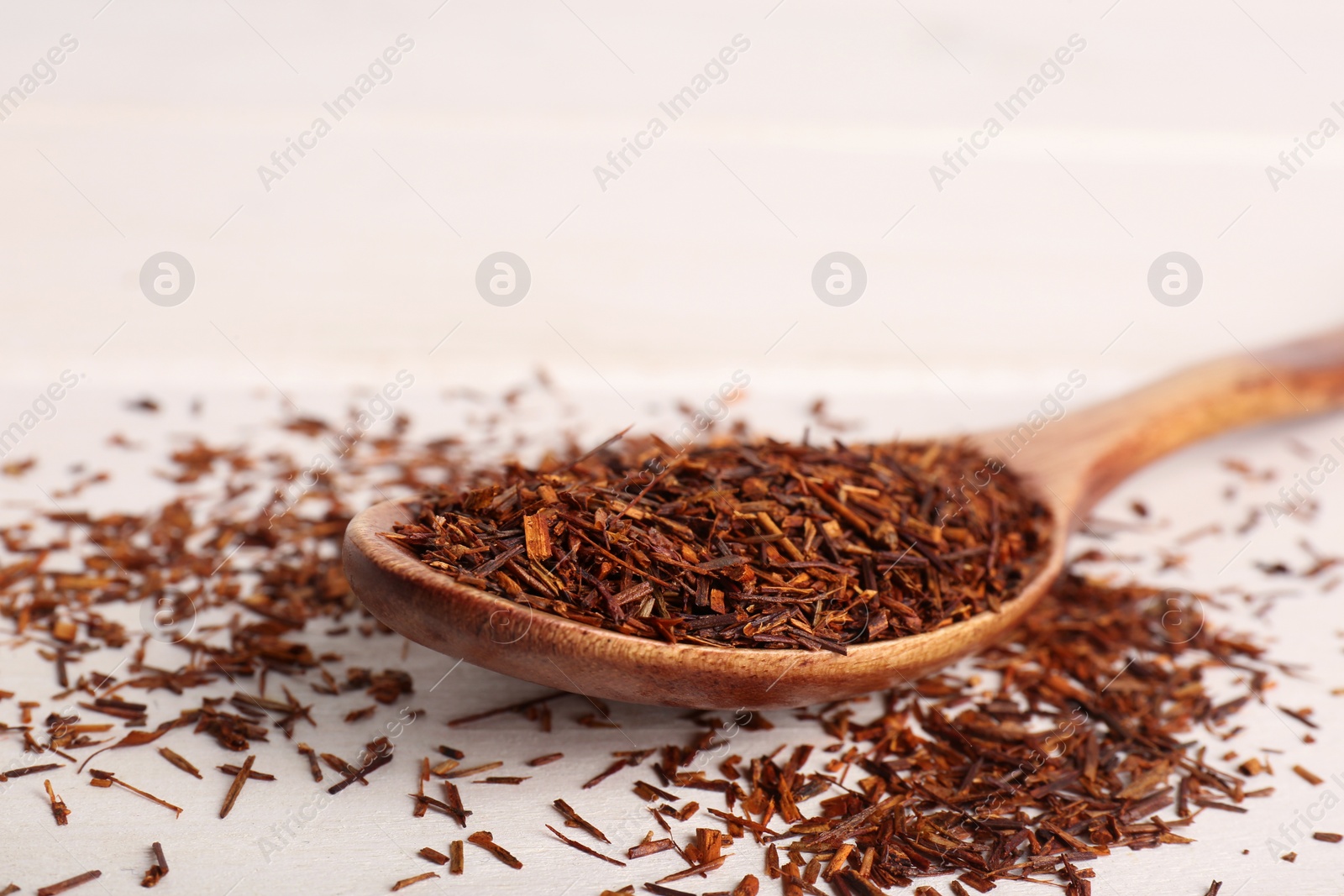 Photo of Spoon with dry rooibos tea leaves on white wooden table, closeup