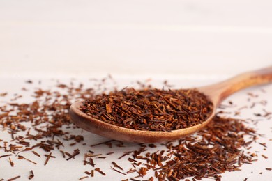 Photo of Spoon with dry rooibos tea leaves on white wooden table, closeup