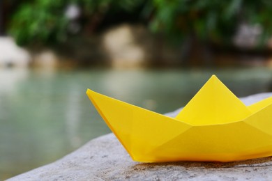 Beautiful yellow paper boat on stone near pond, closeup