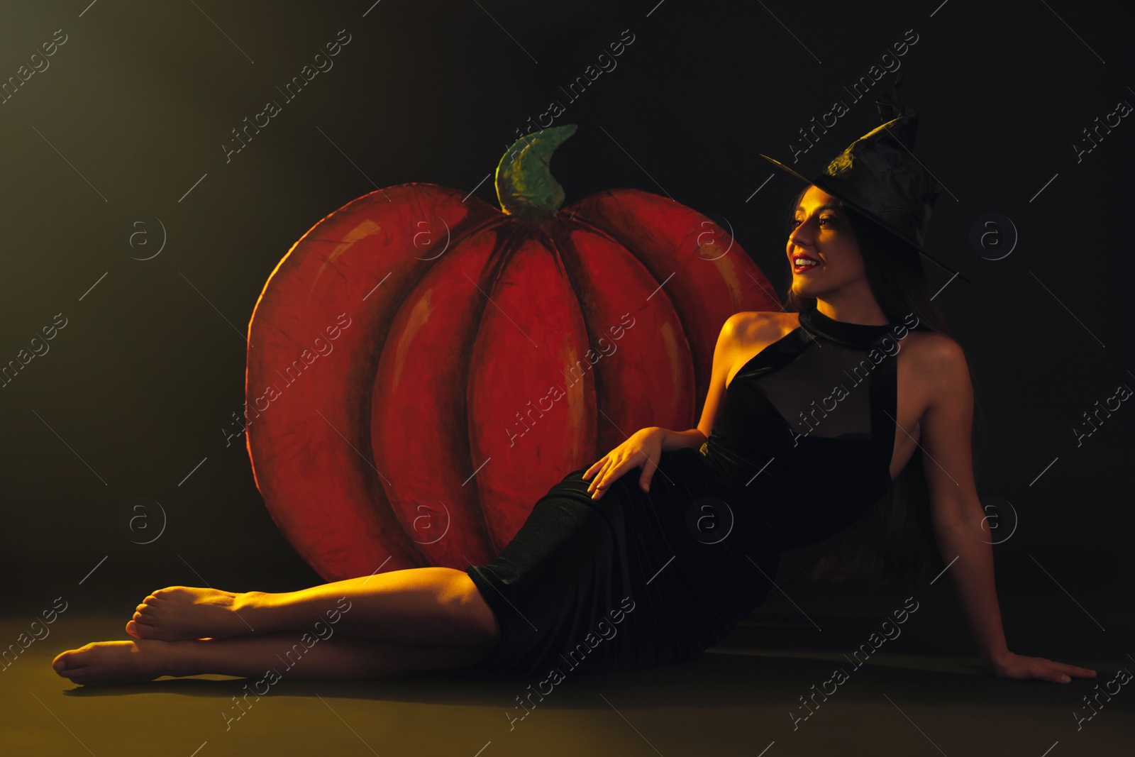 Photo of Young woman wearing witch costume near decorative pumpkin on black background. Halloween party