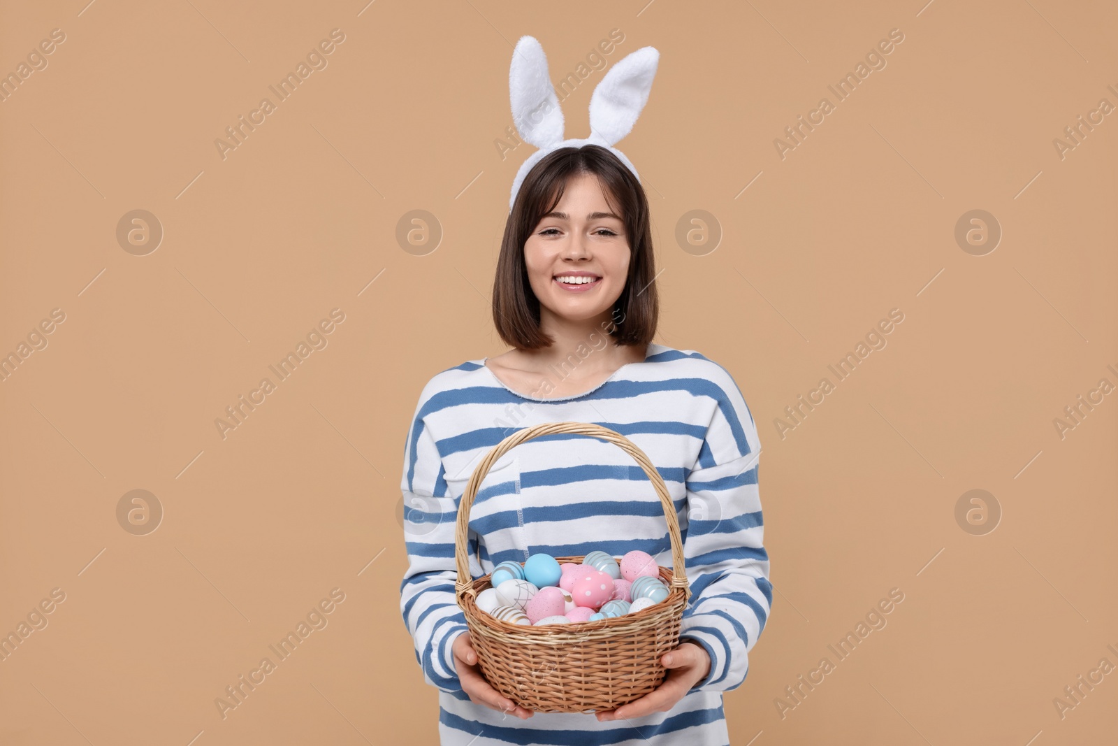 Photo of Easter celebration. Happy woman with bunny ears and wicker basket full of painted eggs on beige background