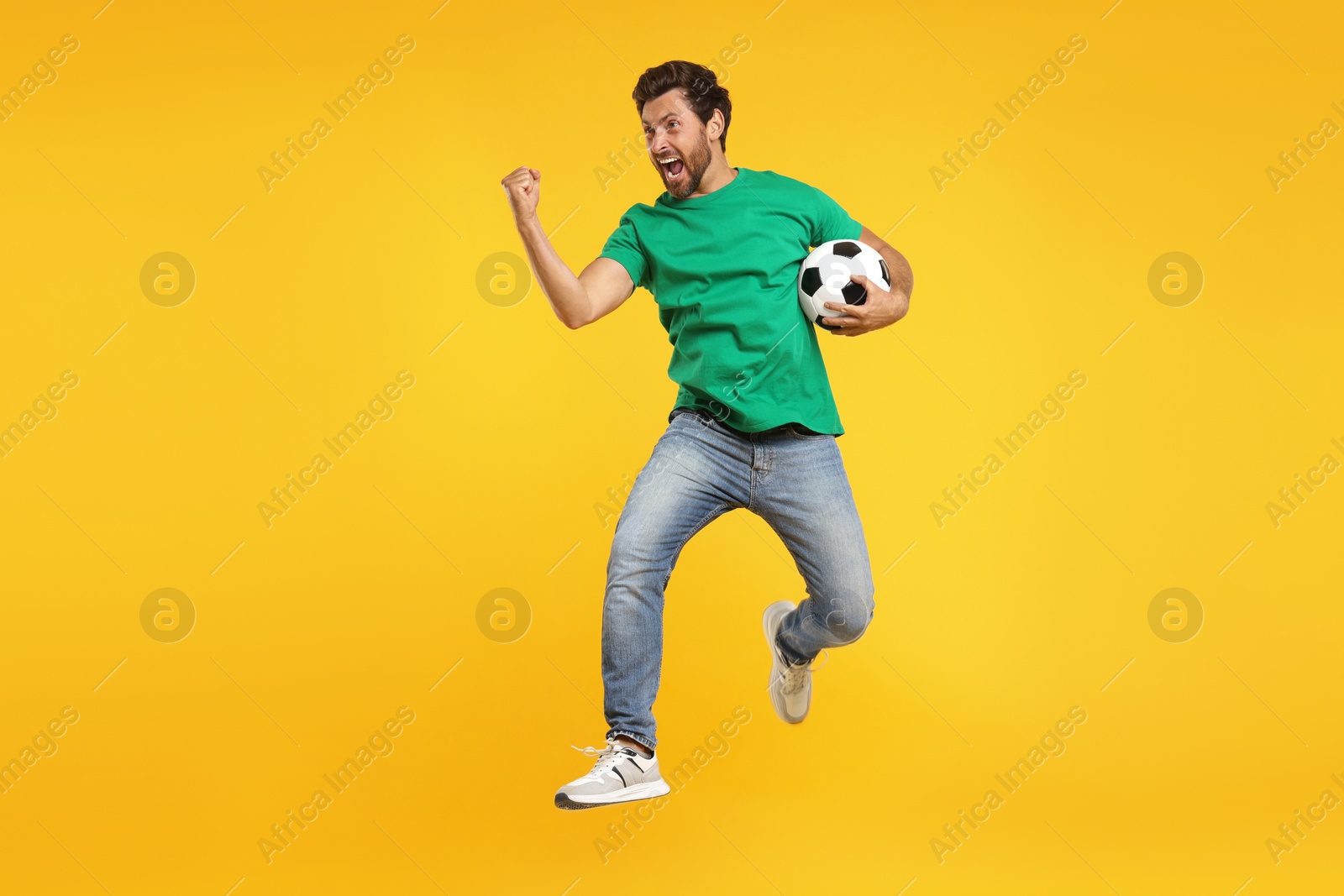 Photo of Emotional sports fan with soccer ball jumping on orange background