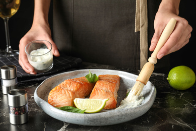 Photo of Woman adding sauce to cooked red fish on dark marble table, closeup