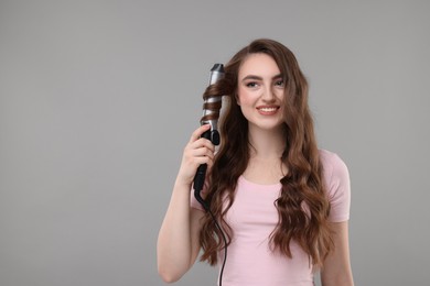 Photo of Beautiful young woman using curling hair iron on grey background, space for text