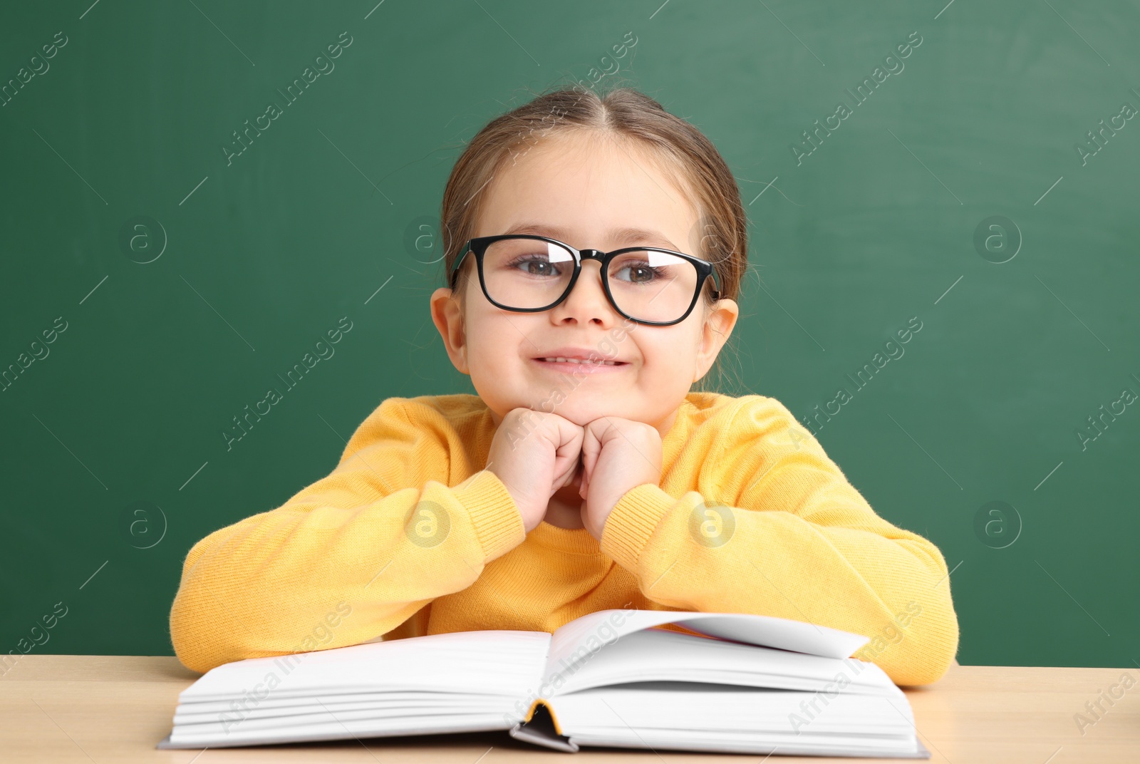 Photo of Happy little school child sitting at desk with book near chalkboard