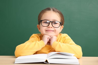 Happy little school child sitting at desk with book near chalkboard