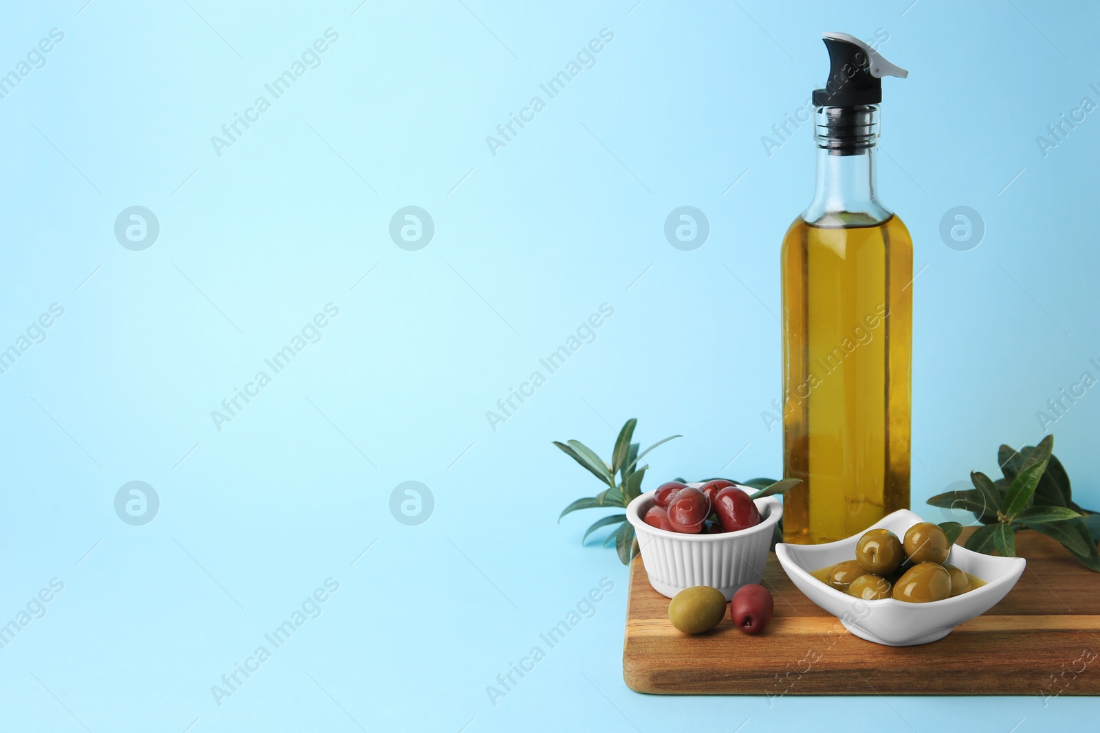 Photo of Bottle of oil, olives and tree twigs on light blue background, space for text