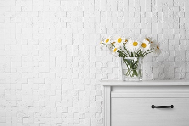 Photo of Beautiful tender chamomile flowers in vase on wooden commode near white textured wall, space for text