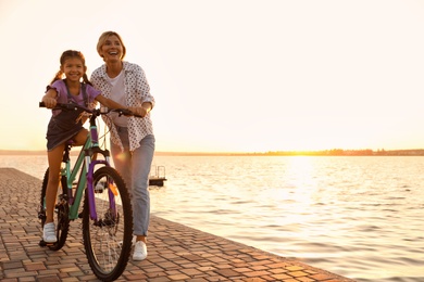 Happy mother teaching her daughter to ride bicycle near river at sunset