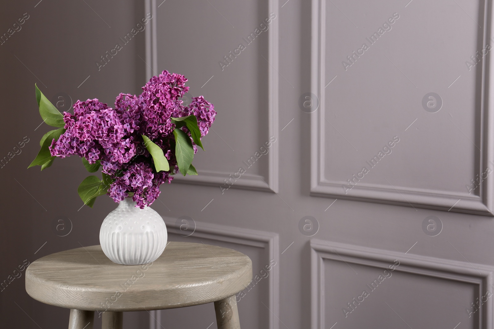 Photo of Beautiful lilac flowers in vase on wooden table near light wall, space for text