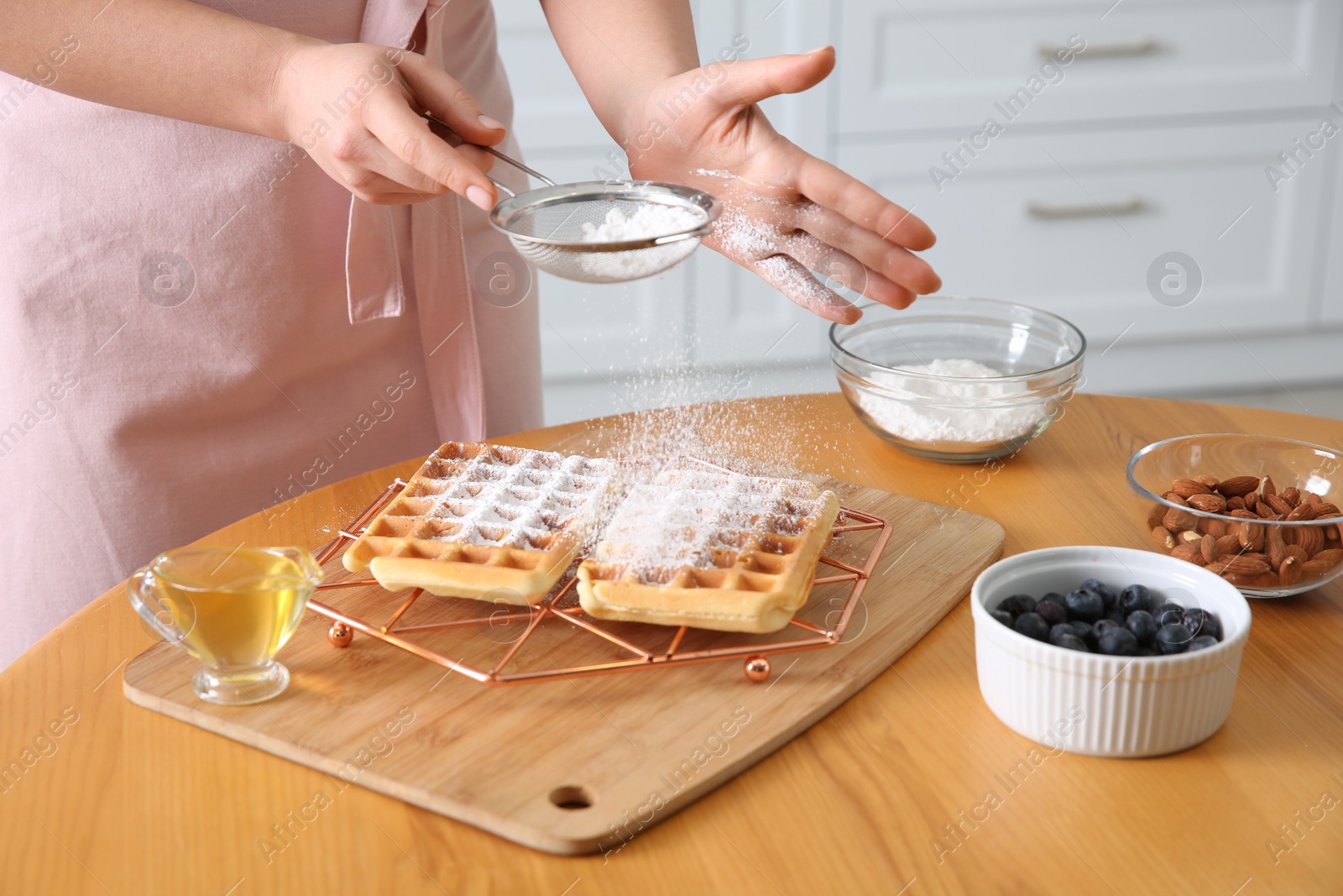 Photo of Woman decorating delicious Belgian waffles with powdered sugar at wooden table in kitchen, closeup