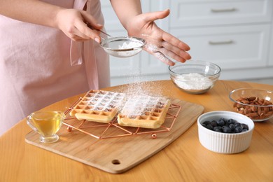 Woman decorating delicious Belgian waffles with powdered sugar at wooden table in kitchen, closeup