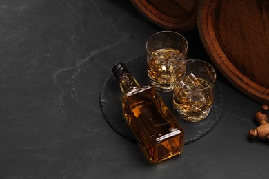 Photo of Whiskey with ice cubes in glasses, bottle and wooden barrels on black table, space for text