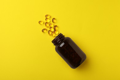 Overturned bottle with dietary supplement capsules on yellow background, flat lay