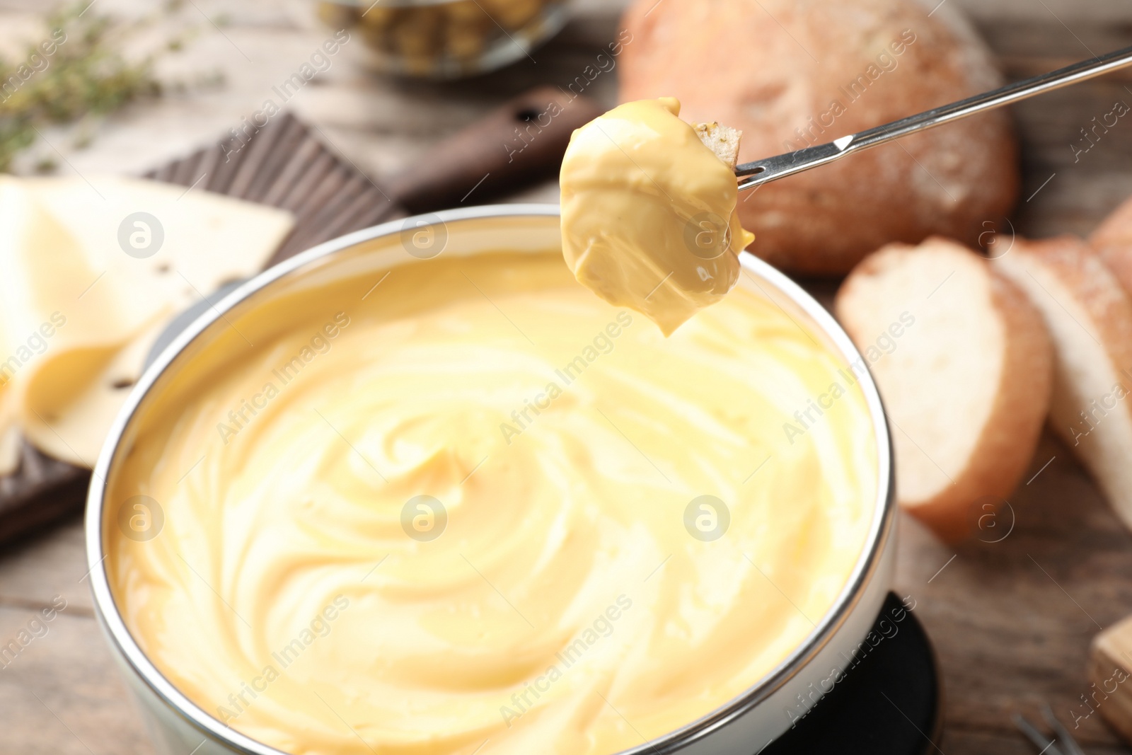 Photo of Piece of bread over pot with delicious cheese fondue on table