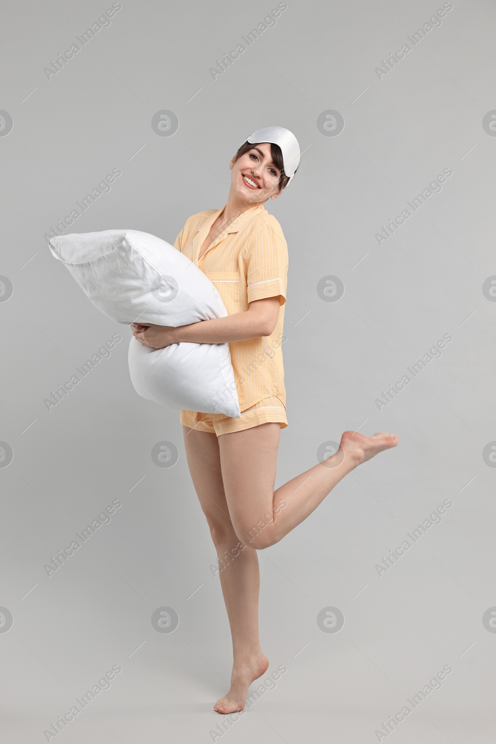 Photo of Happy woman in pyjama and sleep mask holding pillow on grey background
