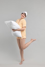 Happy woman in pyjama and sleep mask holding pillow on grey background