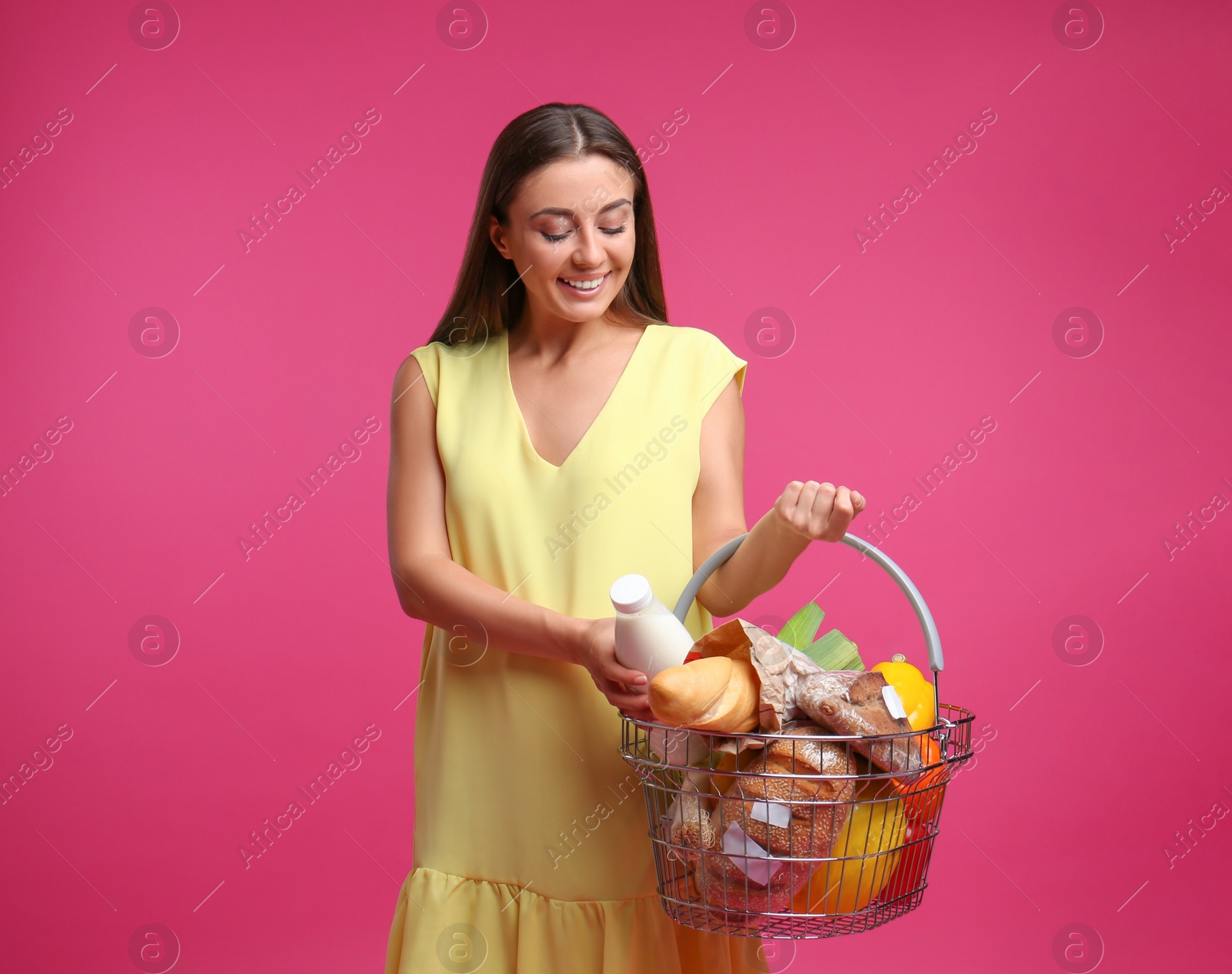Photo of Young woman with shopping basket full of products on pink background