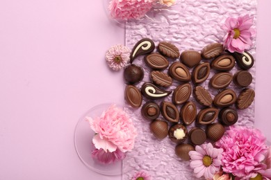 Heart made with delicious chocolate candies and beautiful flowers on pink background, flat lay. Space for text