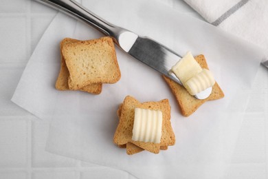 Photo of Tasty butter curls, knife and pieces of dry bread on white tiled table, flat lay