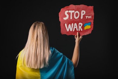 Woman wrapped in Ukrainian flag and holding poster with words Stop the War on black background, back view