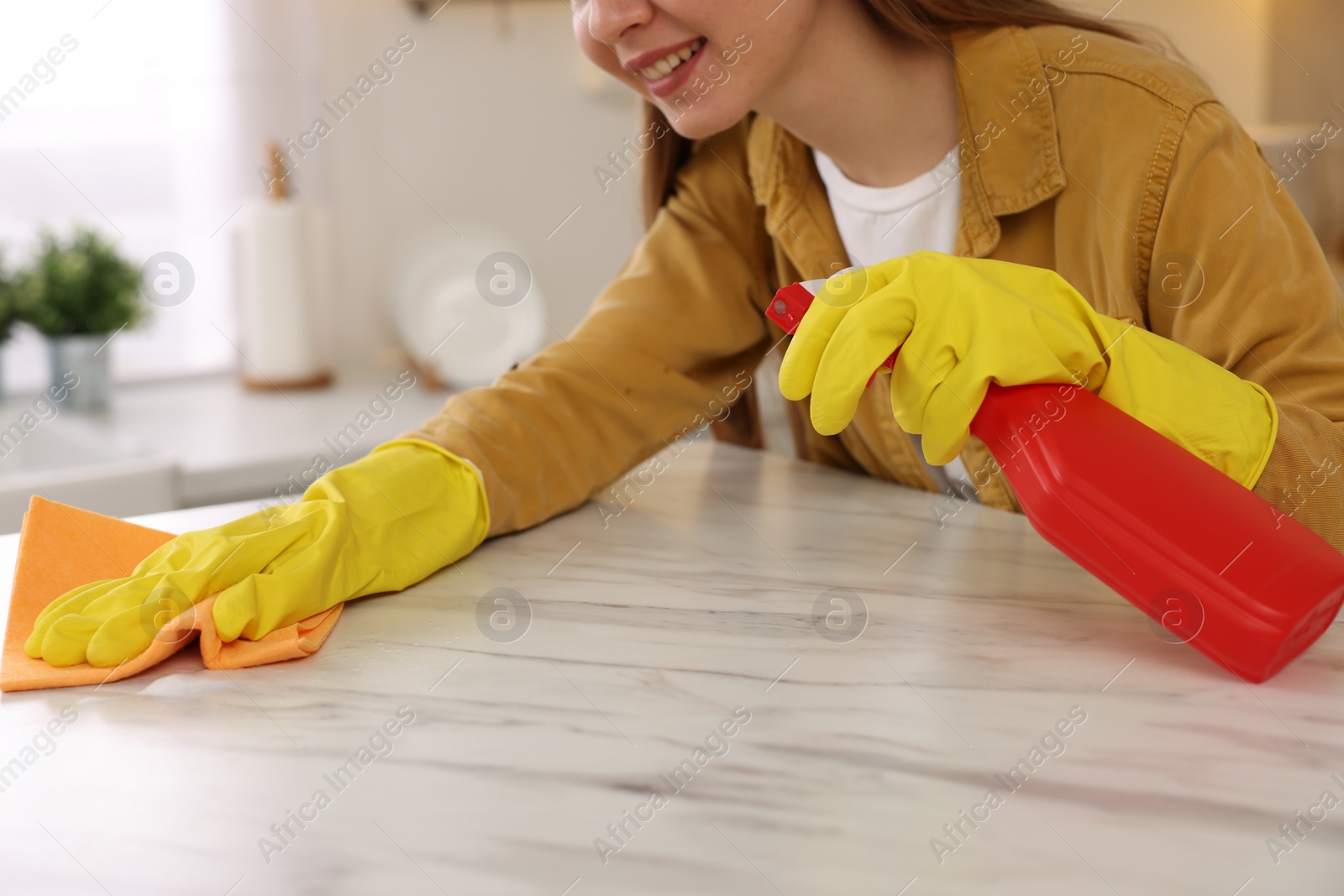 Photo of Woman with spray bottle and microfiber cloth cleaning white marble table in kitchen, closeup