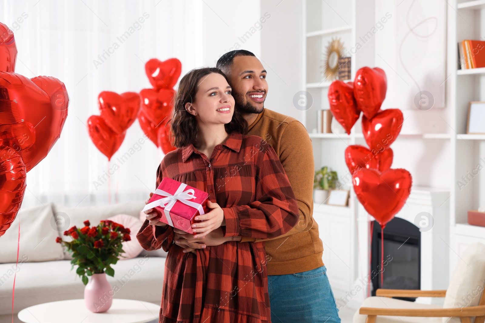 Photo of Happy couple celebrating Valentine's day. Beloved woman with gift box in room decorated with heart shaped air balloons