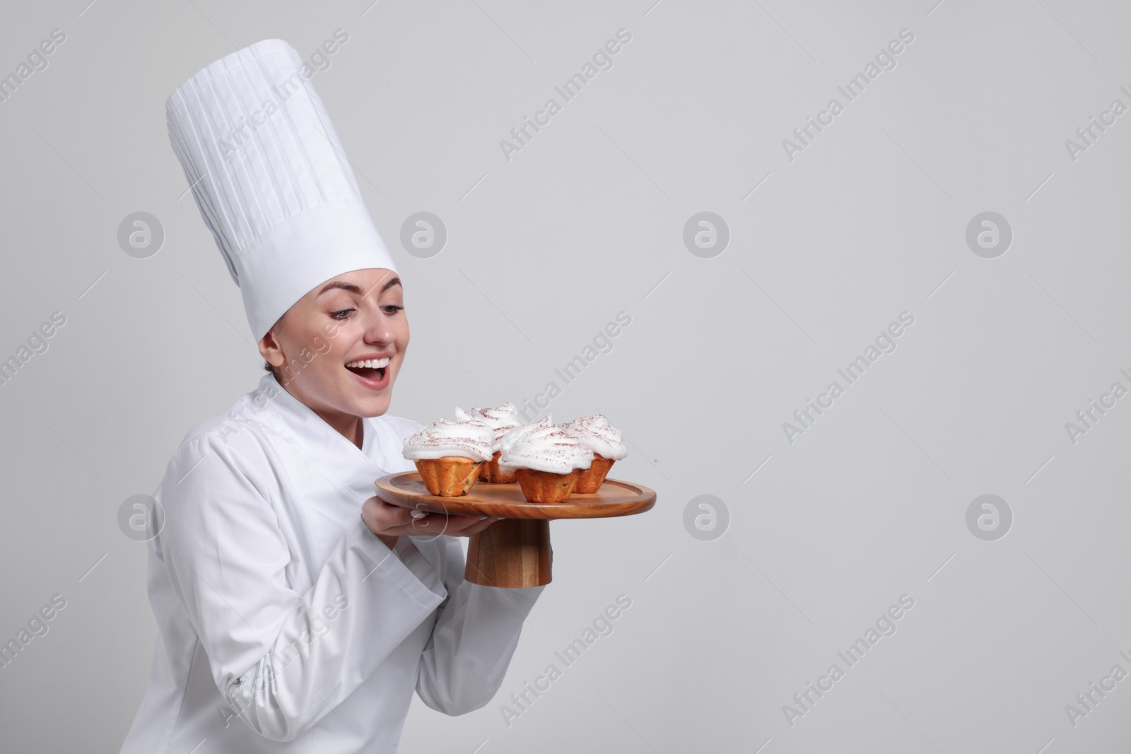 Photo of Happy professional confectioner in uniform holding delicious cupcakes on light grey background. Space for text