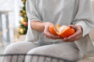 Photo of Woman peeling tangerine in room decorated for Christmas, closeup