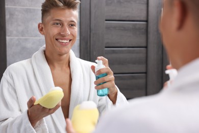 Photo of Happy young man applying face cleanser on sponge near mirror in bathroom