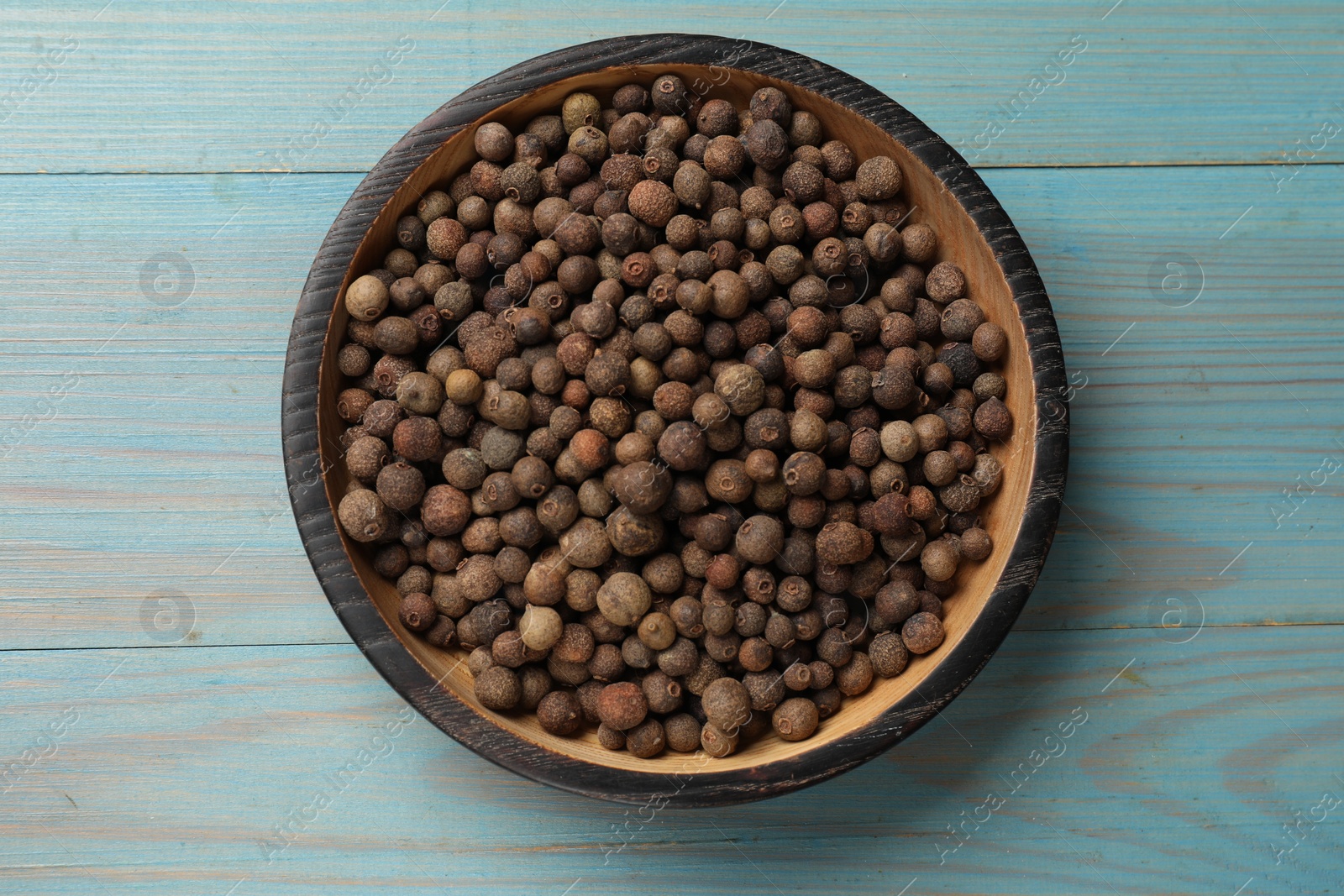 Photo of Aromatic allspice pepper grains in bowl on light blue wooden table, top view
