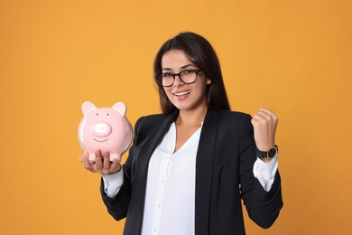 Happy young woman in eyeglasses with piggy bank on orange background